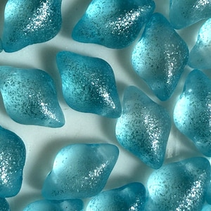 Może przedstawiać: Zbliżenie na liczne, jasnoniebieskie szklane elementy. Elementy mają nieregularny, zaokrąglony kształt i matową fakturę. Szkło ma półprzezroczystą jakość. Elementy są rozrzucone na białej powierzchni, przypominając szkło morskie.