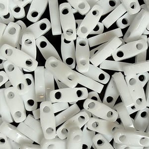 Peut inclure: Gros plan sur une pile de bloque-cordons en plastique blanc. Chaque bloque-cordon est rectangulaire avec des bords arrondis et deux trous pour l'insertion du cordon. Les bloque-cordons sont éparpillés, créant une surface texturée.