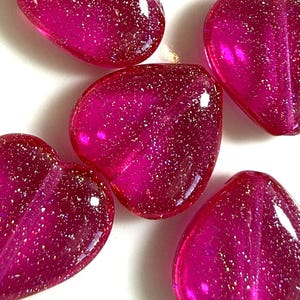 May include: Five translucent, heart-shaped beads in a vibrant magenta hue. Each bead is infused with sparkling glitter, catching the light. The beads are arranged on a white surface, showcasing their glossy finish. Ideal for jewelry making or craft projects.