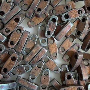 Può includere: Una collezione di piccole perline metalliche rettangolari con una finitura color bronzo anticato. Ogni perlina ha due fori circolari per l'infilatura. Le perline sono sparse, mostrando variazioni di colore e consistenza, adatte per la creazione di gioielli o lavori artigianali.