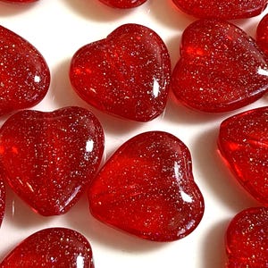 May include: A collection of red, heart-shaped glass beads with glitter embedded throughout. The beads are scattered on a white surface, with varying sizes and shapes. The translucent glass catches the light, creating a sparkling effect.