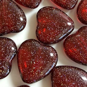 May include: A collection of heart-shaped glass beads in a deep red color, speckled with iridescent glitter. The beads are arranged on a white surface, with their glossy surfaces reflecting light. These decorative beads are suitable for crafts.