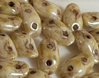 Puede incluir: Una colección de cuentas de semillas de vidrio de color blanco roto con motas marrones y un acabado brillante. Las cuentas tienen forma de grano de arroz y tienen un agujero en el centro.