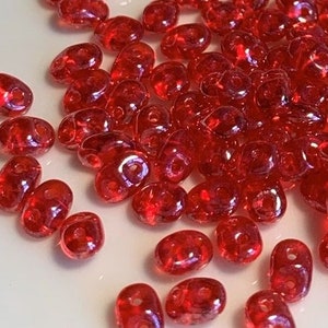 May include: A pile of small, red, iridescent glass seed beads. The beads are oval shaped and have a shiny, reflective surface.