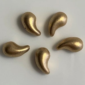May include: Five gold teardrop-shaped beads, arranged in a starburst pattern on a white surface.