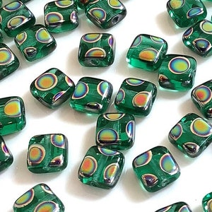May include: A collection of square, emerald green glass beads with iridescent, circular patterns. The beads have rounded corners and a metallic silver lining, scattered across a white surface. These beads are ideal for jewelry making and crafting.