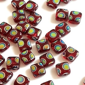 May include: A collection of square, red glass beads with iridescent, multi-colored dot patterns. The beads are scattered across a white surface, showcasing their glossy finish and unique design. Ideal for jewelry making or crafting.