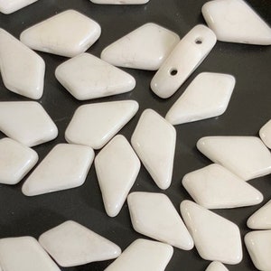 May include: A collection of white, diamond-shaped glass beads. The beads are arranged in a random pattern on a black surface.