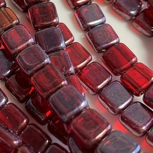 May include: Close-up of strands of square, dark red beads. The beads have a slightly translucent quality, with variations in color and texture. The beads are arranged in parallel lines, creating a visually appealing pattern. Ideal for jewelry making.