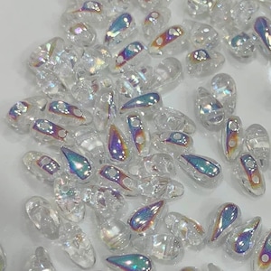 May include: A large quantity of iridescent clear glass teardrop beads. The beads have a pearlescent finish and are scattered on a white surface.