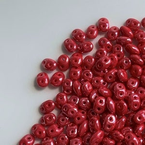May include: A pile of small, red, double-holed beads. The beads have a glossy finish and are oval-shaped, with some being more elongated. They are scattered on a white surface, suggesting a craft or jewelry-making theme.