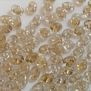 May include: A large quantity of small, clear glass beads with a light gold iridescent sheen. The beads are oval shaped with a hole in the center.