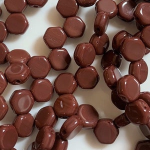 Può includere: Un filo di perline esagonali di vetro marrone. Le perline sono disposte in un mucchio sciolto su una superficie bianca.