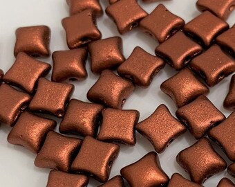 Puede incluir: Un gran grupo de pequeñas cuentas cuadradas de color cobre con un diseño en forma de estrella ligeramente elevado. Las cuentas están esparcidas sobre una superficie blanca.
