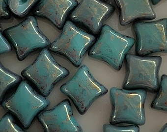 Puede incluir: Una colección de cuentas de azulejos de vidrio azul turquesa con un brillo metálico. Las cuentas tienen forma de cuadrado con esquinas redondeadas.