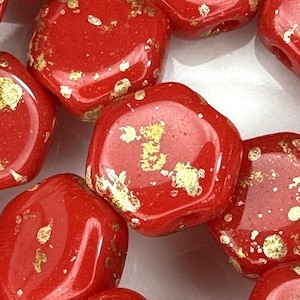 May include: Red hexagonal glass beads with gold flecks. The beads are scattered on a white surface.