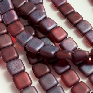 Peut inclure: Gros plan sur plusieurs rangées de perles carrées bordeaux et violettes. Les perles ont une finition mate et sont enfilées. Chaque perle mesure environ 0,6 cm de côté, parfaites pour la création de bijoux.