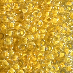 May include: Close-up of a pile of small, transparent yellow seed beads. The beads are scattered, creating a textured surface. The beads are round with a small hole in the center, perfect for stringing. Ideal for jewelry making and crafts.