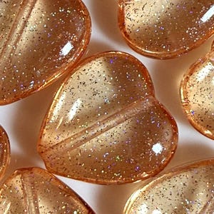 May include: Close-up of several translucent, heart-shaped beads with a glittery, speckled appearance. The beads are a warm, amber color and have a smooth, glossy finish. The beads are likely for jewelry making or craft projects.