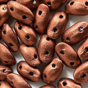 Pode incluir: Uma coleção de contas de vidro de cor cobre com acabamento fosco. As contas são de forma oval e têm dois furos em cada conta.