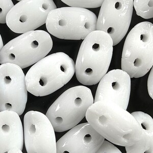 May include: A close-up of a group of white oval-shaped beads with two holes each. The beads are smooth and glossy.