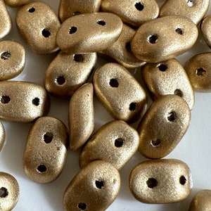 Op de afbeelding: Een groep gouden, ovale kralen met elk twee gaten. De kralen hebben een matte afwerking en zijn verspreid over een wit oppervlak.