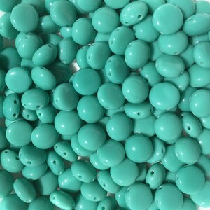 May include: A large group of turquoise blue glass beads. The beads are oval shaped and have two holes in each bead.