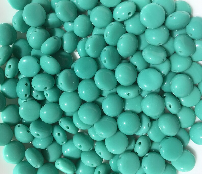 May include: A large group of turquoise blue glass beads. The beads are oval shaped and have two holes in each bead.