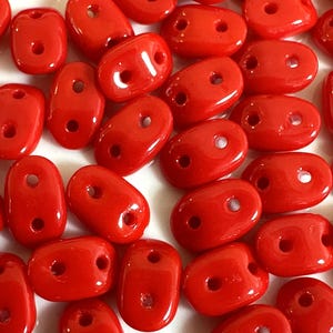 May include: A close-up of a group of bright red oval-shaped beads with two holes each. The beads are scattered on a white surface.