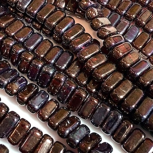 May include: Close-up of strands of rectangular beads in shades of brown, purple, and bronze. The beads have a glossy, polished finish and are strung together, creating a textured surface. The beads are likely for jewelry making.