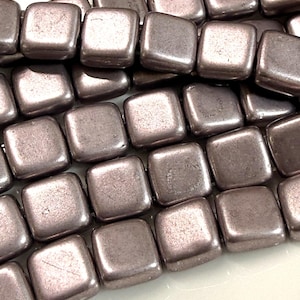 May include: Close-up of a strand of square, metallic beads in a muted mauve color. The beads have a slightly textured surface and rounded edges. The beads are strung together, creating a continuous line of the square shapes. Ideal for jewelry making.