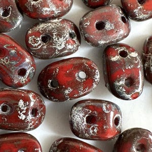 May include: A group of red and silver oval-shaped beads with two holes each. The beads have a speckled, textured surface.