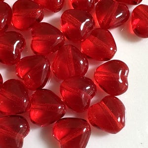 May include: A collection of translucent red heart-shaped beads. Each bead has a diagonal stripe across its center. The beads are scattered, with some overlapping, creating a textured and romantic display. Ideal for jewelry making or craft projects.