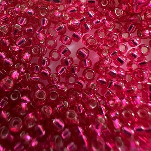 May include: Close-up of a pile of small, translucent, hot pink seed beads. The beads have a metallic, iridescent finish and are cylindrical with a hole through the center. These beads are ideal for jewelry making and other craft projects.