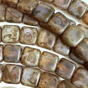 May include: Close-up of square beads in shades of brown, gold, and lavender. The beads have a slightly iridescent finish and are strung together. The beads are likely for jewelry making or other craft projects.