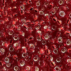 May include: A close-up shot of numerous small, transparent red glass beads. Each bead has a hole running through the center and a metallic gold-colored lining. The beads are scattered, creating a textured, reflective surface.