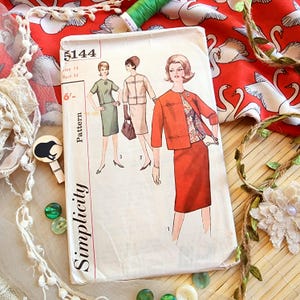 May include: A vintage Simplicity sewing pattern, number 5144, showing illustrations of women's outfits. The envelope is cream-coloured with red text and illustrations. The background includes red fabric with white swan designs and green thread.