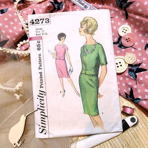 May include: A vintage Simplicity sewing pattern, number 4273, with illustrations of a green and pink dress. The pattern is for a junior size 11 with a 80 cm bust. The image includes buttons, fabric, and a wooden kiwi bird.
