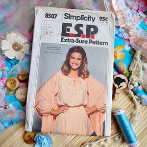 May include: A vintage Simplicity ESP Extra-Sure Pattern sewing pattern, size H, featuring a woman in a peach-coloured dress. The pattern includes Time Saver stretch knit methods. The image is surrounded by fabric, buttons, and thread.