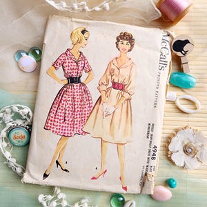 Peut inclure: Un patron de couture vintage McCall's avec des illustrations de deux femmes en robes chemisiers. L'une est imprimée de cœurs, l'autre est de couleur unie avec une ceinture. Des accessoires de couture et des éléments décoratifs sont disposés autour.