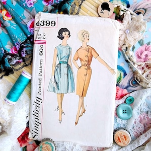 Pode incluir: Padrão de costura Simplicity vintage, número 4399, tamanho 12, com ilustrações de dois vestidos. Um é um vestido listrado azul claro, o outro um vestido bege. O fundo inclui tecido floral e acessórios de costura.