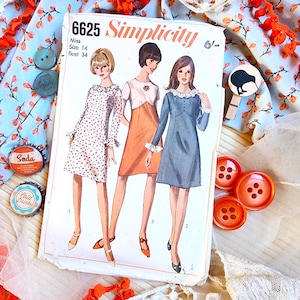 Op de afbeelding: Vintage Simplicity naaipatroon, maat 14, met drie jurk ontwerpen. De omslag toont illustraties van de jurken in verschillende kleuren. De afbeelding bevat oranje knopen, frisdrankflesdoppen en bloemenstof.