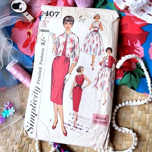 May include: Vintage Simplicity sewing pattern featuring illustrations of dresses and a jacket. The pattern cover is cream-colored with red and white floral accents. The text "Simplicity Printed Pattern" is visible, along with the number "2407".