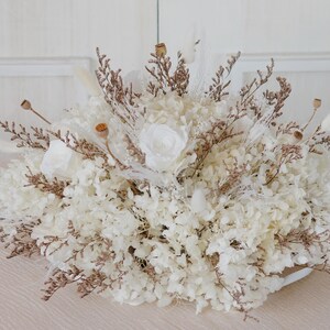 May include: A circular floral arrangement composed of white hydrangeas, roses, and dried flowers, with brown and beige accents. The arrangement is a natural, elegant design, suitable for home decor or special events.