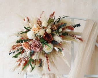 May include: A bouquet of dried flowers with pampas grass, roses, and other dried foliage. The bouquet is arranged on a white metal arch with a sheer fabric draped over it.