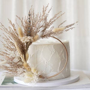 May include: A white frosted cake with a gold ring and dried floral arrangement. The arrangement includes beige pampas grass, white flowers, and small white bead-like accents. The cake sits on a white cake stand.