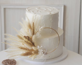 May include: Two-tiered white wedding cake with a gold ring topper and a bouquet of dried pampas grass and white flowers.