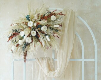 May include: A white metal archway decorated with a bouquet of dried pampas grass, white roses, and eucalyptus. A sheer white fabric drapes over the archway.