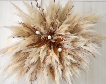 May include: A large, fluffy wreath made of dried pampas grass and other dried grasses. The wreath is decorated with small white pom poms.