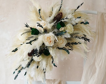 May include: A floral arrangement featuring cream-colored pampas grass, white roses, and dark green accents. Pinecones and sprigs of greenery are interspersed throughout the bouquet. The arrangement is set against a neutral background.